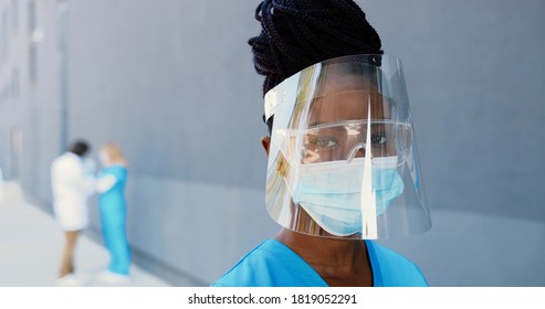 Portrait African American Beautiful Woman Doctor In Medical Mask, Face Shield And Goggles Looking At Camera. Close Up Female Physician In Respiratory Protection. Doctors On Background. Protected Suit.