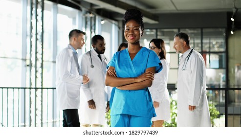 Portrait Of African American Beautiful Woman Medic Smiling. Turning Face To Camera And Standing In Clinic. Indoors. Pretty Female Doctor Or Nurse In Hospital. Multi Ethnic Docs Talking On Background.