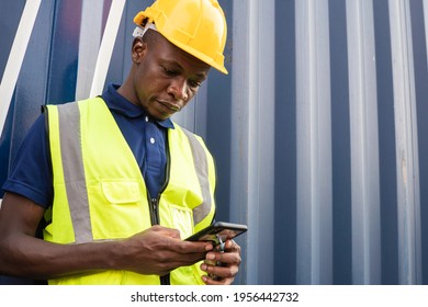 Portrait Of African American Black Male Factory Worker In Safety Uniform Checking Work, Playing Smartphone, Online Chatting Or Browsing On Mobile Phone While Taking A Break At Construction Site