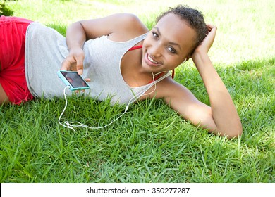 Portrait Of An African American Black Teenager Girl Laying On Green Grass In A Park, Relaxing Listening To Music Using A Smartphone And Headphones, Smiling Outdoors. Adolescent Technology Lifestyle.