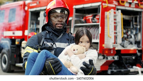 Portrait Of African American Brave Fireman Holds Saved Girl In His Arms Standing Near Fire Truck. Firefighter In Fire Fighting Operation