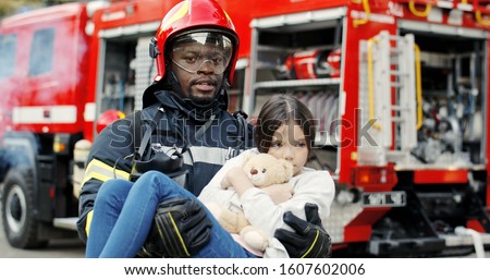 Portrait of african american brave fireman holds saved girl in his arms standing near fire truck. Firefighter in fire fighting operation