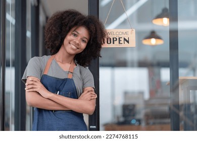 Portrait of African American businesswoman standing at the cafeteria door entrance. A cheerful young waitress in a blue apron near a glass door with an open signboard and looking at the camera. - Powered by Shutterstock