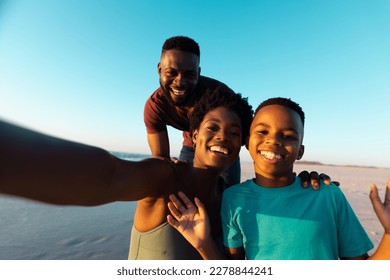 Portrait of african american cheerful woman taking selfie with husband and son at beach against sky. Copy space, sunset, unaltered, family, together, childhood, nature, vacation, enjoy, technology. - Powered by Shutterstock