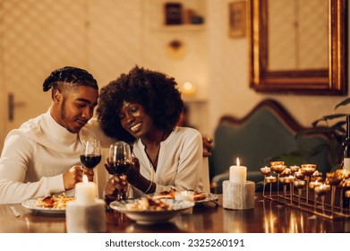 Portrait of an african american couple having a romantic candlelight dinner at home and watching a video on a smartphone together. Concept of love and relationship. Black spouses using a mobile phone. - Powered by Shutterstock