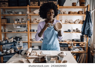 Portrait of an african american craftswoman at pottery studio on workshop and decorating earthenware while having wine. Smiling multicultural pottery craftswoman doing her hobby and having wine. - Powered by Shutterstock