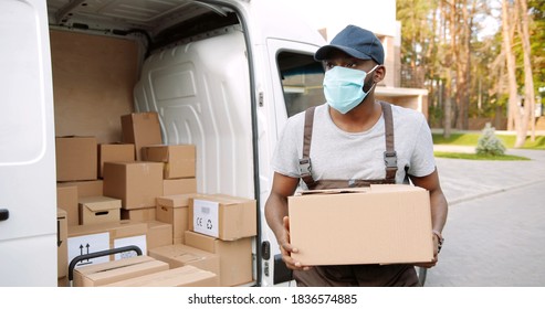 Portrait Of African American Delivery Man In Mask Holding Carton Box In Front Of His Van. 