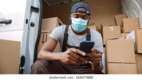 Portrait Of African American Delivery Man In Mask And Uniform  Using Smartphone While Sitting In His Van. 