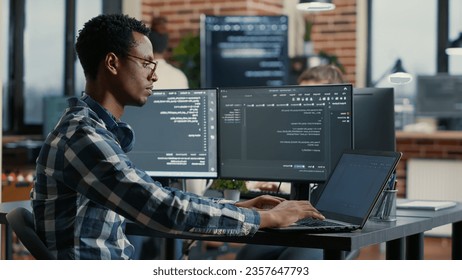 Portrait of african american developer using laptop to write code sitting at desk with multiple screens parsing algorithm in software agency. Coder working on user interface using portable computer.