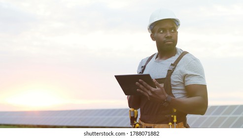 Portrait Of African American Electrician Engineer In Safety Helmet And Uniform Using Tablet Checking Solar Panels. 