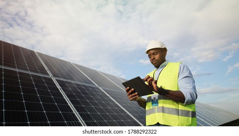 Portrait of african american electrician engineer in safety helmet and uniform using tablet checking solar panels. 