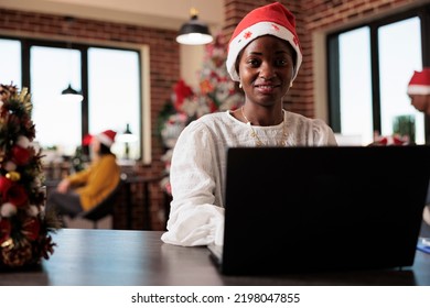 Portrait of african american employee wearing santa hat, working on business at company office filled with christmas decorations and tree lights. Using laptop in startup workplace with xmas decor. - Powered by Shutterstock