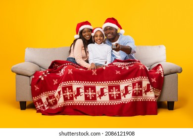 Portrait Of African American Family Of Three People Watching Christmas Movies On TV Sitting On Sofa At Home Covered With Red Blanket, Enjoying Xmas Eve Holidays Television Show, Yellow Orange Wall - Powered by Shutterstock