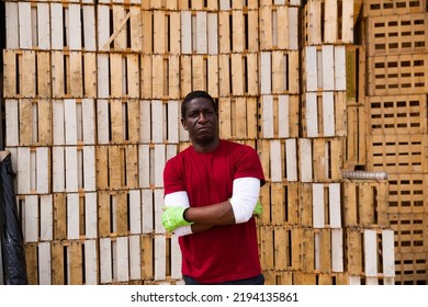 Portrait Of African American Farm Worker Standing With Crossed Arms On Background With Stack Of Used Wooden Crates