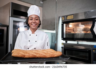 Portrait of African American female chef in white cooking uniform looking at camera with cheerful smile and proud with tray of baguette in kitchen, pastry foods professional, fresh bakery occupation. - Powered by Shutterstock