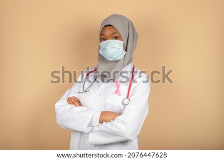 Portrait of an African American female doctor wearing a surgical mask, scarf and white coat posing with her arms crossed.