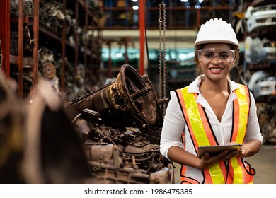 Portrait Of African American Female Worker In Safety Vest Working With Digital Tablet And Checking Old Automotive Spare Parts, Engine For Maintenance In Automotive Spare Parts Storage Warehouse