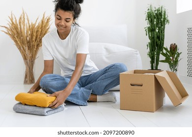 Portrait of african american girl putting pullovers into big box in comfortable room with decorations. Clothes charity concept. Garment donation. Preparation for moving - Powered by Shutterstock