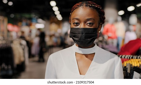 Portrait Of African American Girl Woman Shopper Consumer Female Consultant Salesperson Wears Black Medical Face Mask Looking At Camera Stands In Clothing Store In Shopping Center In Covid Quarantine