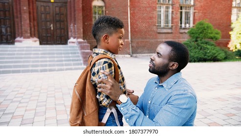 Portrait Of African American Handsome Young Man Talking With Cute Son At Schoolyard Outside. Nice Male Junior Student With School Bag Speaking With Father Near School. Family Concept