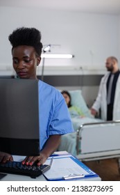 Portrait of african american health care worker using personal computer for reading lab results in hospital ward. Medical doctors giving clinical consult while nurse looks at medical history.