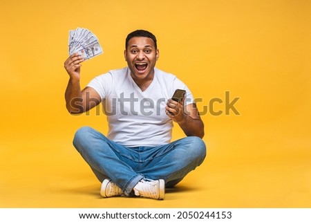 Portrait of african american indian black young man sitting on the floor holding dollar banknotes isolated over yellow background. Using mobile phone.