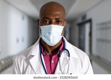 Portrait of african american male doctor wearing lab coat and face mask in corridor at hospital. Hospital, hygiene, medicine, healthcare and work, unaltered. - Powered by Shutterstock