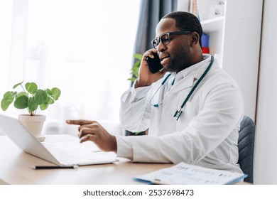 Portrait of African American male doctor in glasses talking on mobile phone looking at laptop screen. Healthcare, medicine concept - Powered by Shutterstock