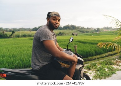Portrait of African American male tourist looking at camera during time for moped trip around Bali in Indonesia, handsome man with helmet resting at motor bicycle during daytime on vacations