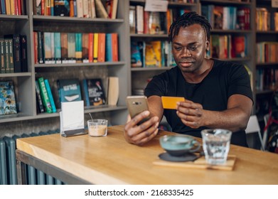 Portrait Of An African American Man Holding Credit Card And A Smartphone Over Wooden Cafe Table With Coffee Cup In A Cafe. Paying Bill At Cafe. Using Mobile Banking Application During Lunch.