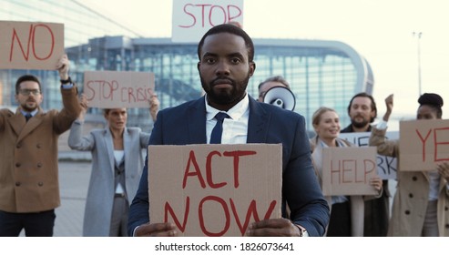 Portrait Of African American Man In Suit And Tie Standing Outdoor At Protest With Poster Act Now. Male Protester At Strike Against Unemployment. Mixed-races People On Background. Protesting. Striking.