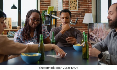 Portrait Of African American Man Thinking About Strategy Play At Card Games With Friends, Having Fun In Living Room. Young Adult Enjoying Board Game Play With Tactics, Drinking Beer With People.