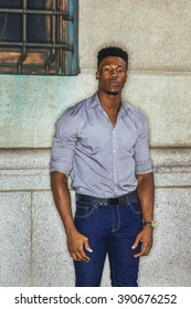 Portrait Of African American Man. Wearing Gray Shirt, Blue Jeans, Wristwatch, Young Black Guy With Little Goatee, Standing Against Wall With Small Window On Street In New York, Looking At You.
