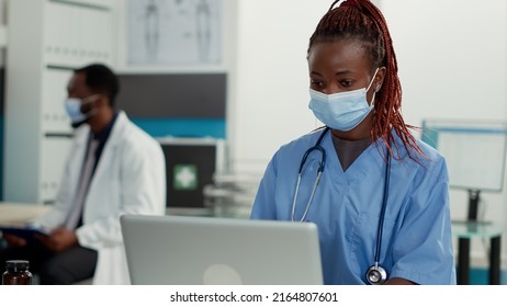 Portrait Of African American Nurse With Face Mask Working On Laptop, Using Technology To Plan Patient Appointments. Medical Assistant Sitting At Cabinet Desk For Health Care Service.