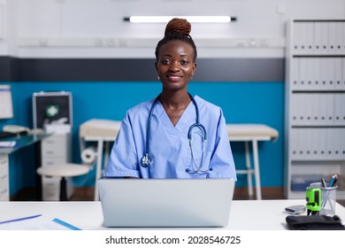 Portrait Of African American Nurse Using Laptop At White Desk Sitting In Medical Office. Black Woman With Assistant Occupation Wearing Uniform And Working At Healthcare Clinic