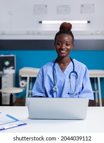 Portrait Of African American Nurse Using Laptop At White Desk Sitting In Medical Office. Black Woman With Assistant Occupation Wearing Uniform And Working At Healthcare Clinic