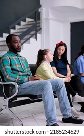 Portrait Of African American Patient Looking Tired Waiting In Busy Hospital Reception Room With Diverse People. Bored Man Sitting Down Looking Pensive Waiting For Receptionist To Call His Name.