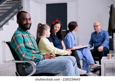 Portrait Of African American Patient Smiling At Camera While Professional Nurse Is Admitting Older Man In Hospital Clinic. Confident Man Sitting Down In Hospital Reception With Diverse People.