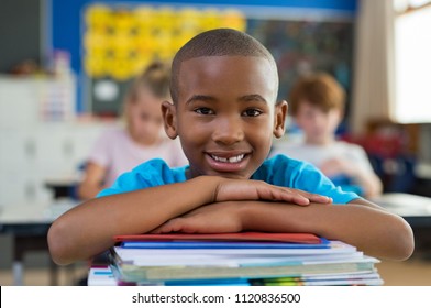 Portrait of african american schoolboy leaning on desk with classmates in background. Happy young kid sitting and leaning chin on stacked books. Portrait of elementary pupil looking at camera. - Powered by Shutterstock