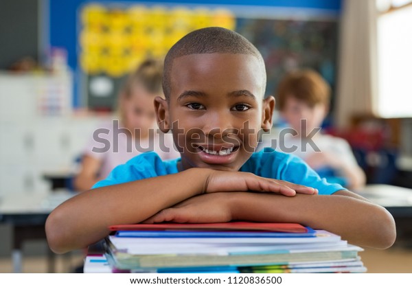Portrait of african american schoolboy leaning on\
desk with classmates in background. Happy young kid sitting and\
leaning chin on stacked books. Portrait of elementary pupil looking\
at camera.