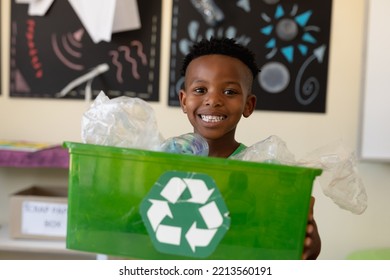 Portrait of an African American schoolboy with short hair holding a green crate with a white recycling logo on it and looking to camera smiling in an elementary school classroom - Powered by Shutterstock