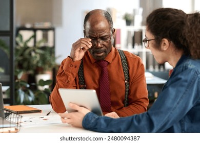 Portrait of African American senior man adjusting glasses while looking at tablet screen in business meeting with young colleague in office copy space - Powered by Shutterstock