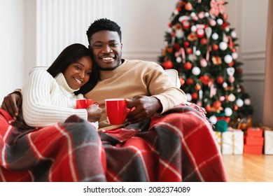 Portrait Of African American Spouses Watching Christmas Movies On TV Drinking Coffee Sitting On Sofa At Home Covered With Blanket. Family Enjoy Xmas Eve Holidays Television Show In Cozy Living Room