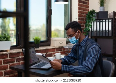 Portrait Of African American Startup Owner With Glasses Wearing Face Mask Focused On Clipboard Analyzing Marketing Indicators. Entrepreneur Comparing Bussiness Data With Sales Results On Laptop.