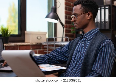 Portrait Of African American Startup Owner With Glasses Analyzing Marketing Indicators On Clipboard With Charts Looking At Laptop. Entrepreneur Comparing Bussiness Data With Sales Results.