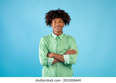 Portrait of african american teen guy with folded arms looking and smiling at camera, wearing shirt, posing isolated over blue studio background. Happy casual male teenage model laughing