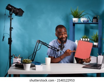 Portrait Of African American Vlogger Doing Product Review Of Red Book Pointing Finger At Mockup Cover In Vlogging Studio. Influencer Sitting At Desk With Podcast Recording Setup Reviewing Novel.