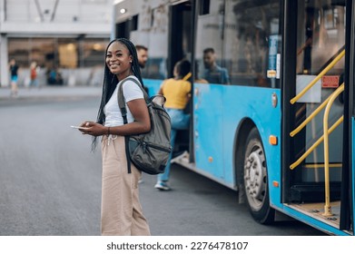 Portrait of an african american woman looking at the camera while waiting at a bus stop. Young black female using public transportation while going to school or work. Using smartphone for bus schedule - Shutterstock ID 2276478107