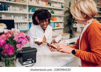 Portrait of an african american woman pharmacist assisting a senior woman customer while using a smartphone and standing at the counter in a pharmacy. Customer Support in a drugstore. Diversty. - Powered by Shutterstock