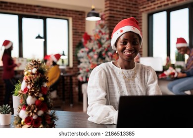 Portrait of african american woman with santa hat working on business at company office filled with christmas decorations and tree lights. Using laptop in startup workplace with xmas decor. - Powered by Shutterstock